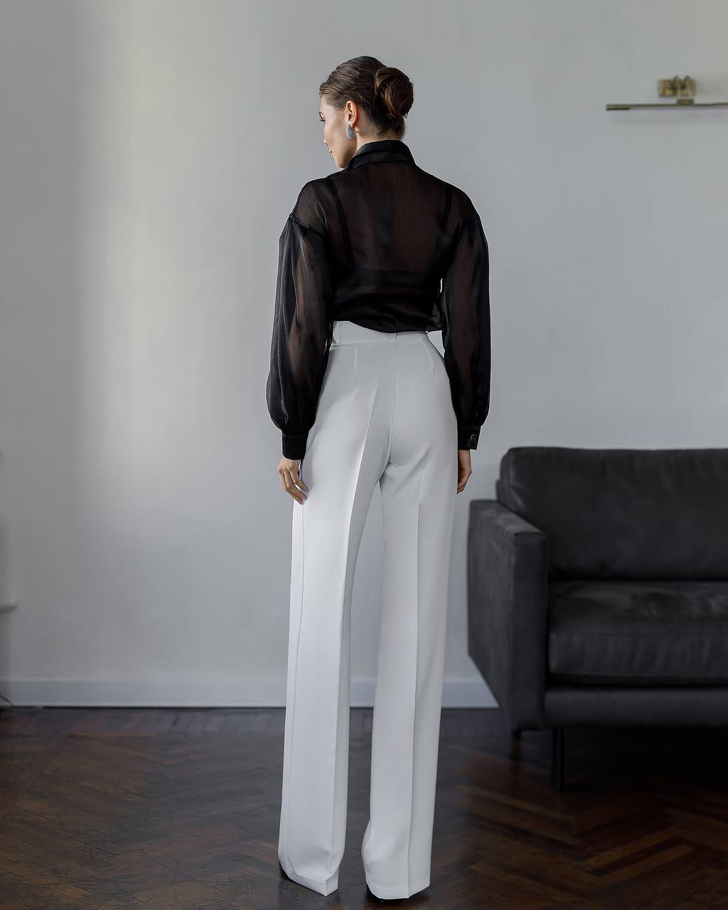 Person wearing a black sheer blouse and white pants standing in a room with a gray sofa.