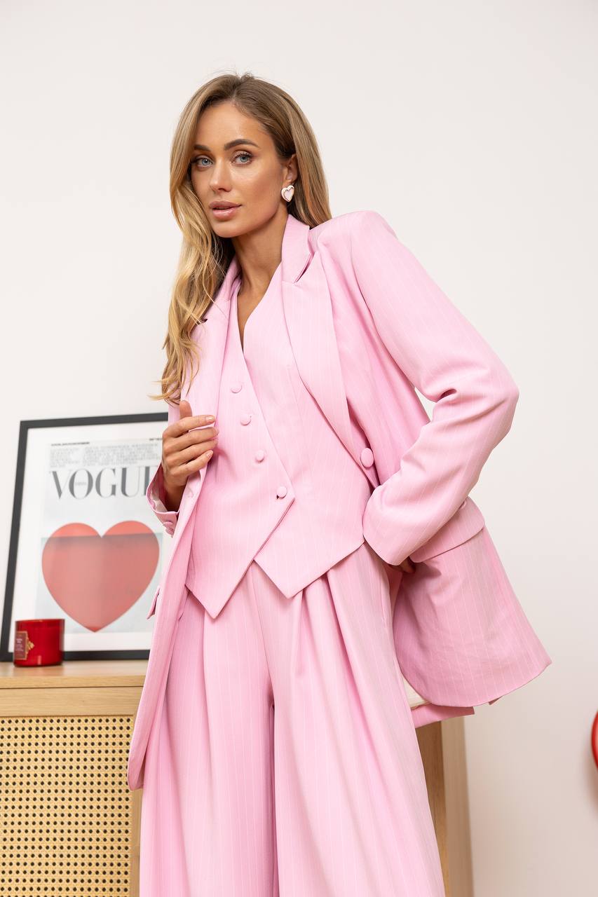 Woman wearing a pink suit standing in front of a white wall with a framed picture of a heart and 'Vogue' magazine.
