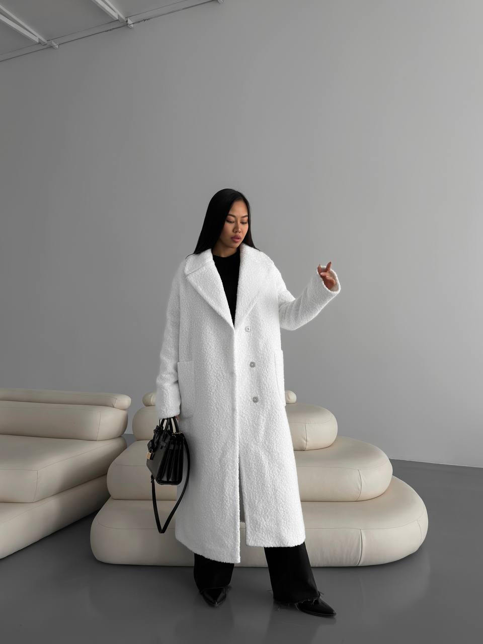 Woman in a white coat standing in a modern interior setting with a gray wall and beige sofa.