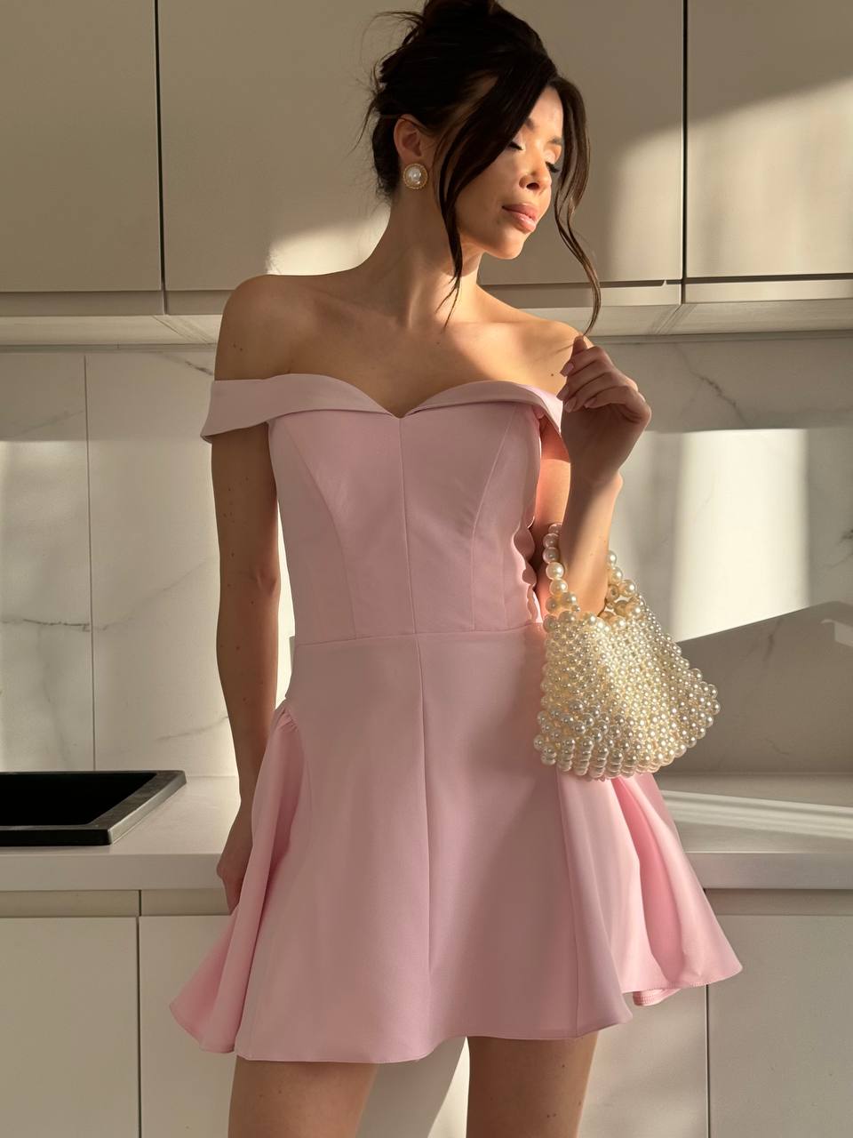 Woman in a pink off-shoulder dress holding a pearl-handled bag in a kitchen.