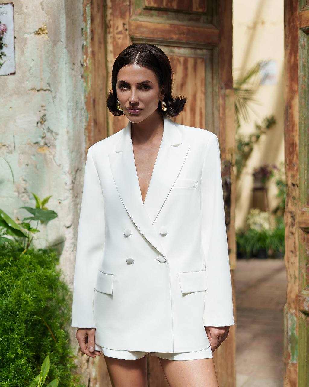Woman wearing a white blazer and shorts set in an outdoor setting with plants and a rustic wall.