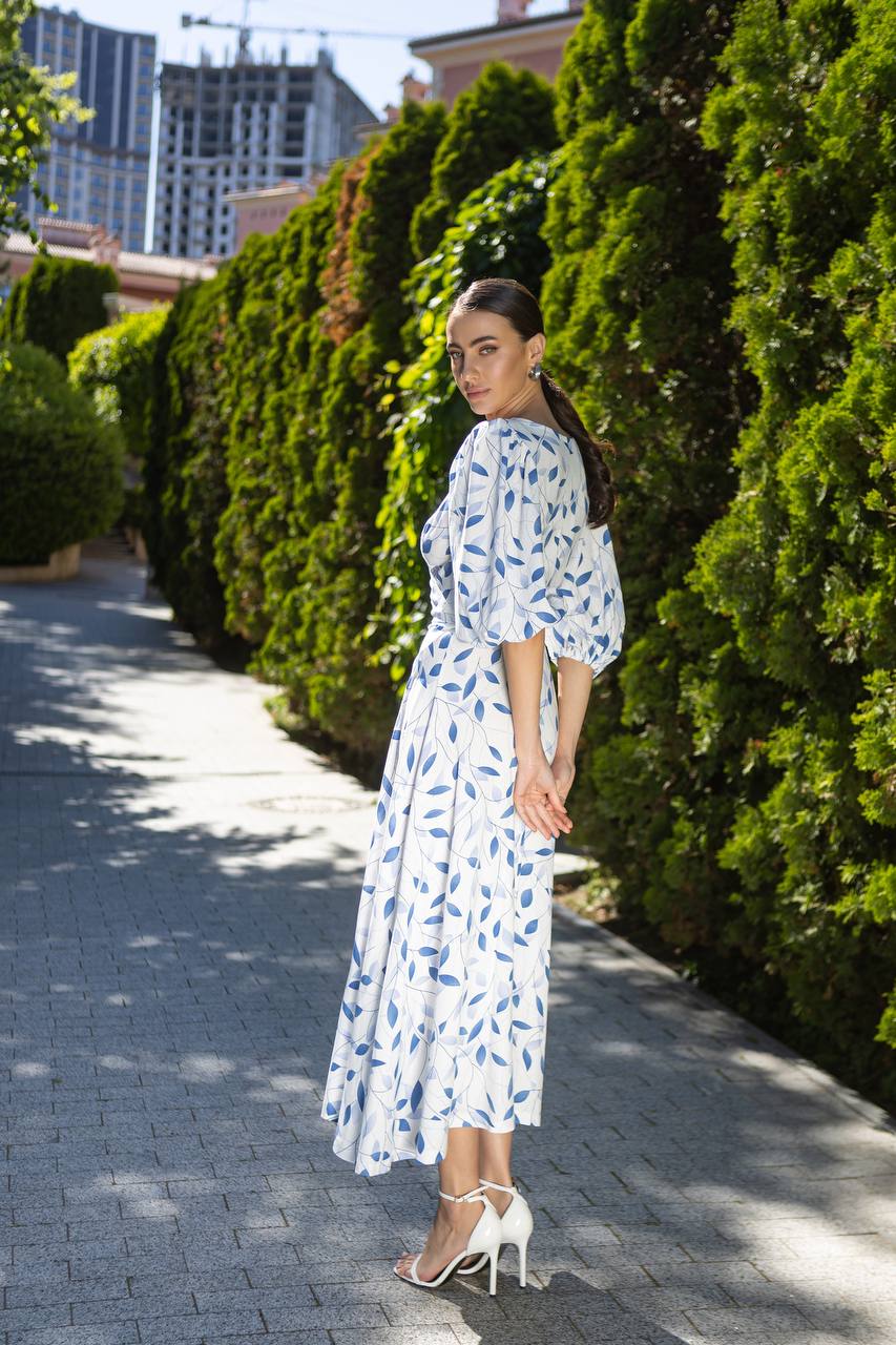 Woman in a white dress with blue patterns walking along a sunlit path lined with green hedges.