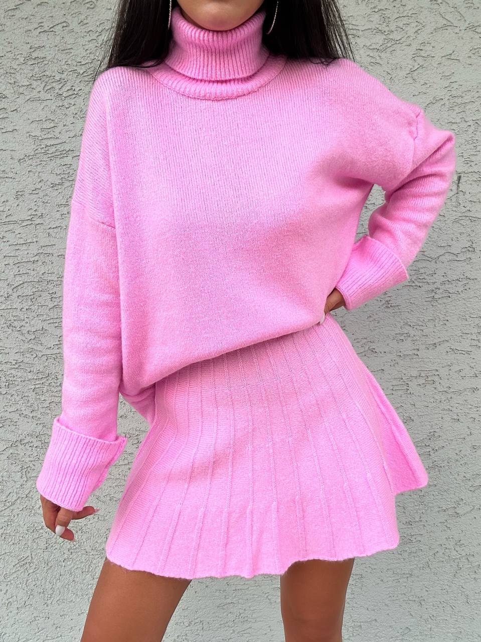 Person wearing a pink ribbed sweater and skirt set against a gray background