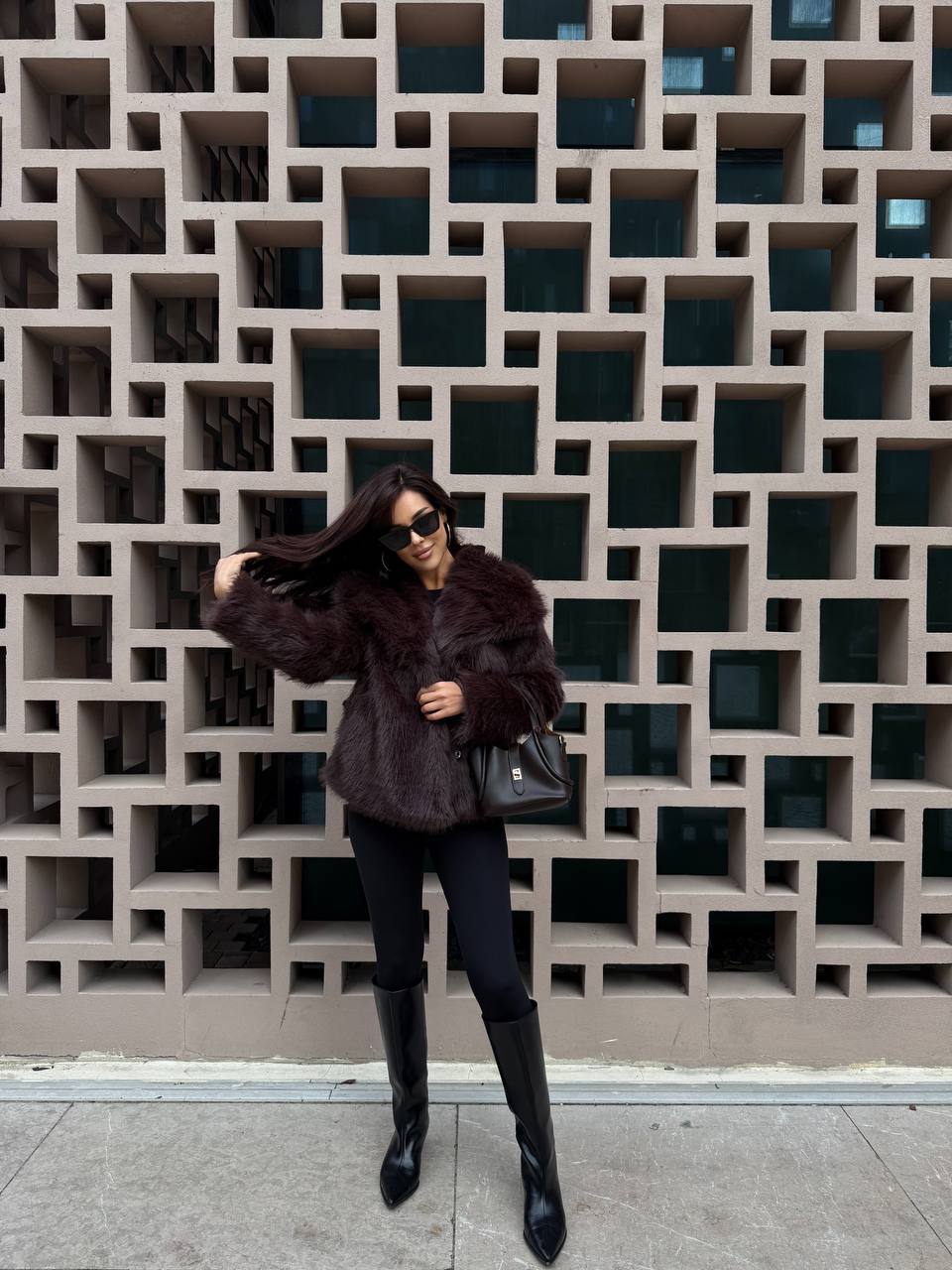 Person wearing a fur coat and black boots standing in front of a geometric wall.