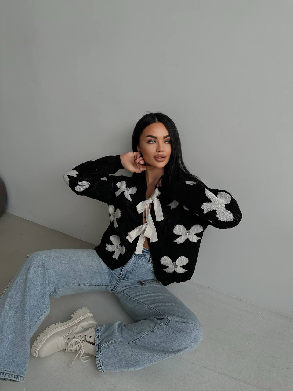 Woman wearing a black cardigan with white floral patterns and blue jeans sitting on a gray floor.