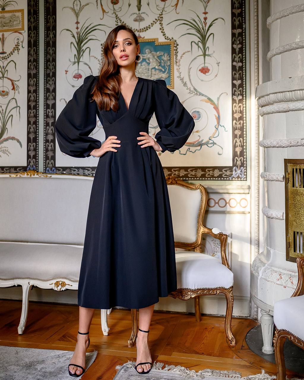 Woman in a black dress standing in an ornate room with decorative walls and furniture.