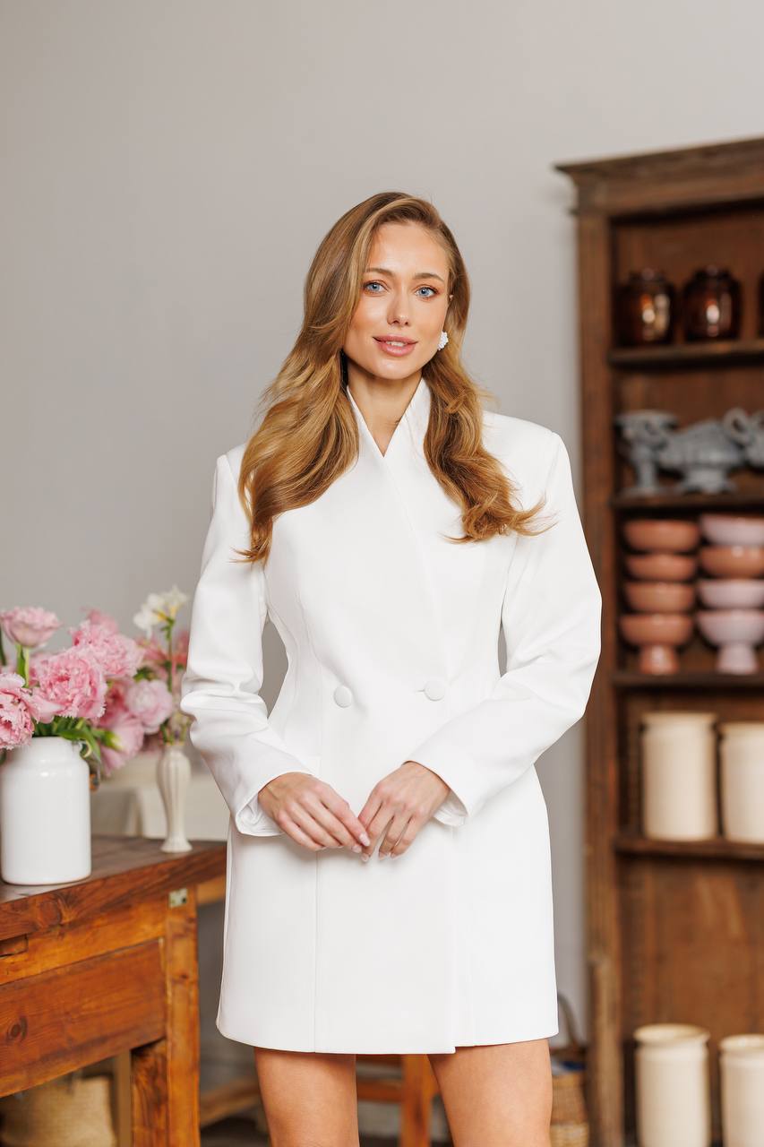Woman in a white coat standing in a room with wooden furniture and decorative items.