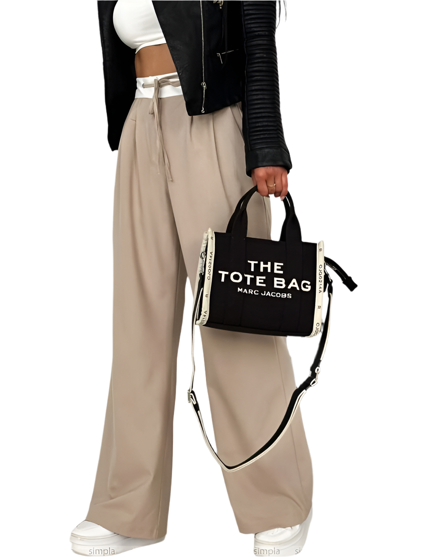 Person holding a black tote bag with 'The Tote Bag' text, wearing beige pants and a black jacket.
