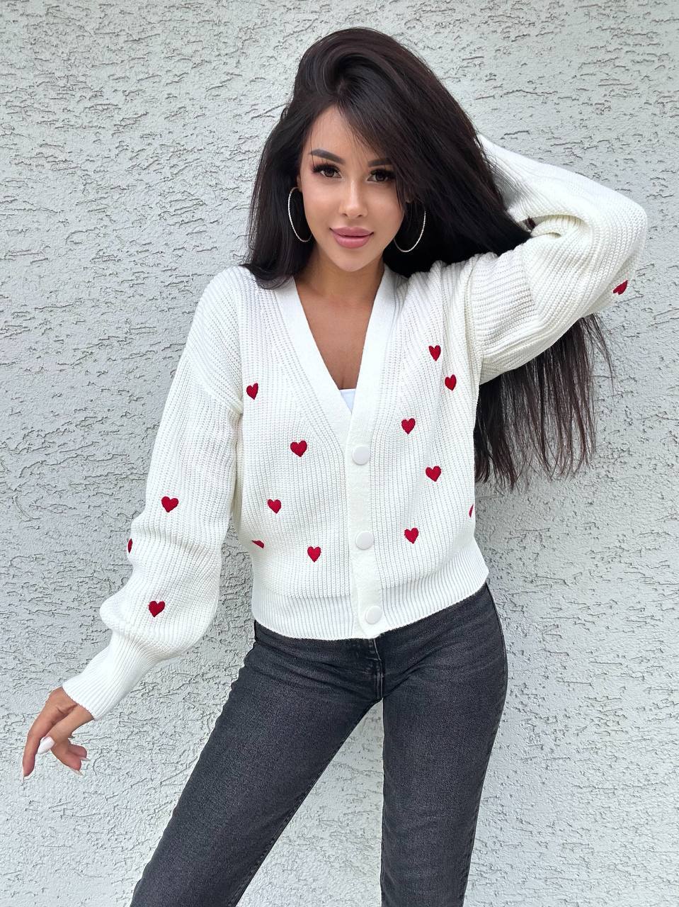 Woman wearing a white sweater with red heart patterns against a gray wall.