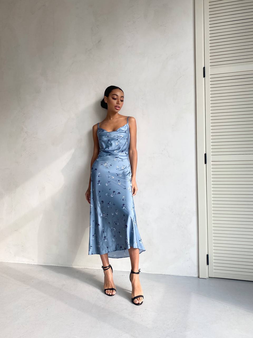 Woman wearing a blue dress with floral patterns against a plain wall.