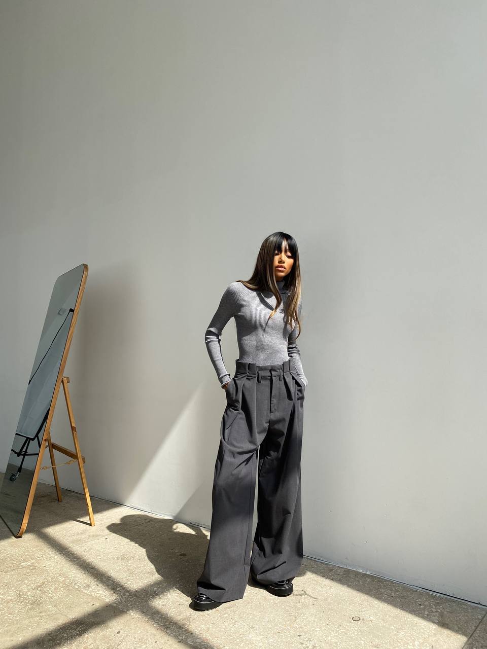 Woman in gray outfit standing against a white wall with a wooden stand and mirror in the background.