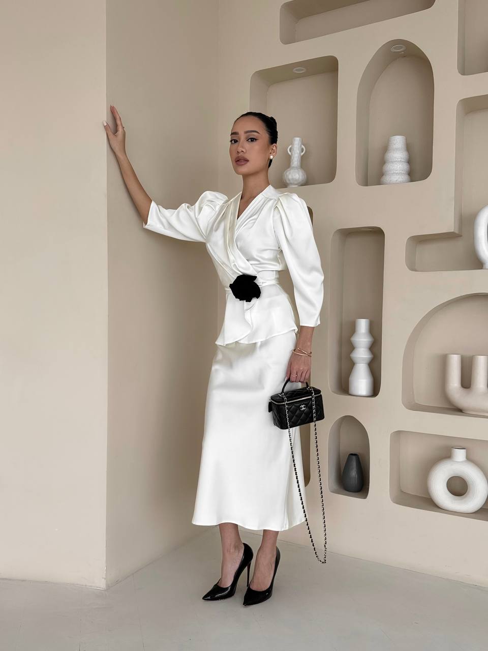 Woman in a white outfit with black accessories standing in a room with decorative shelves.