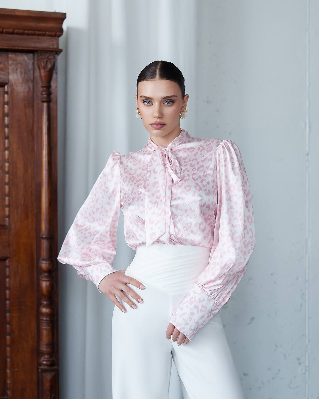 Woman wearing a pink floral blouse and white pants against a light gray wall.