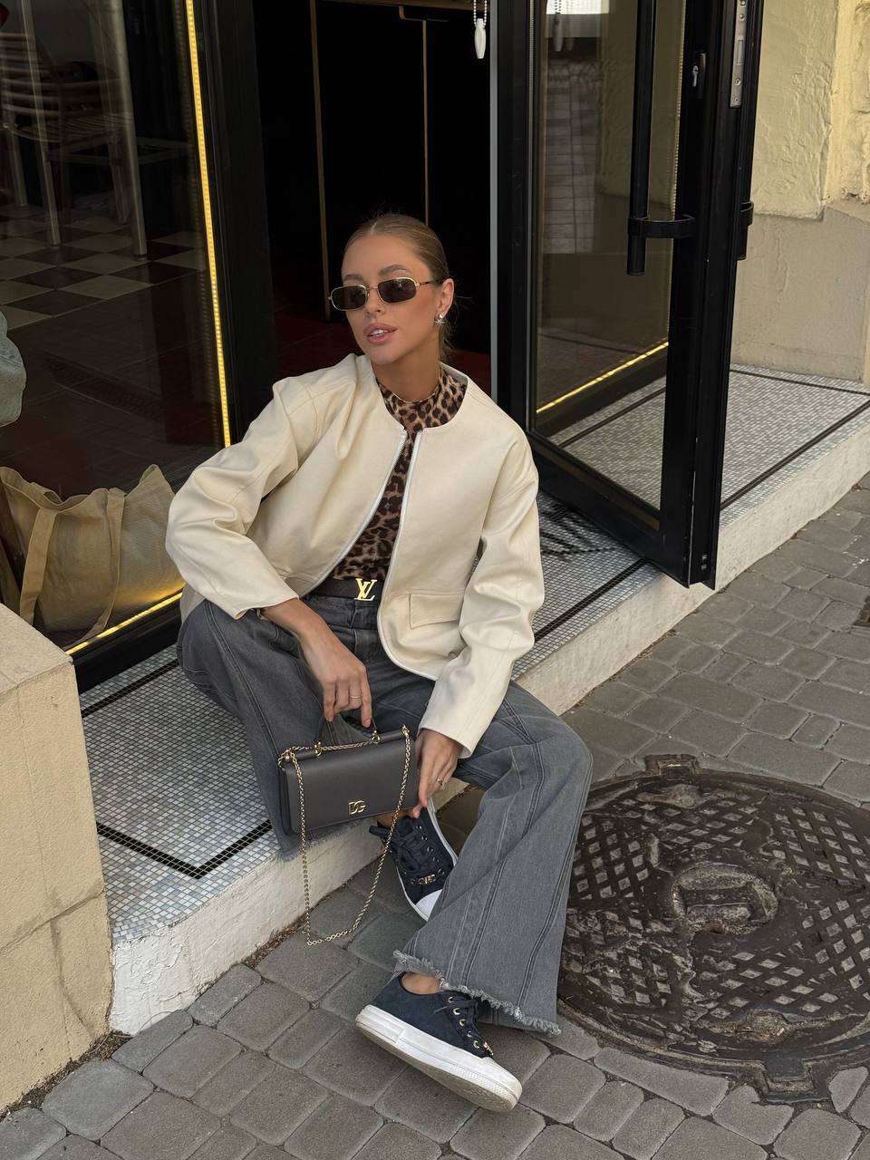 Woman sitting on a step wearing sunglasses, a white jacket, and blue jeans with a black handbag.