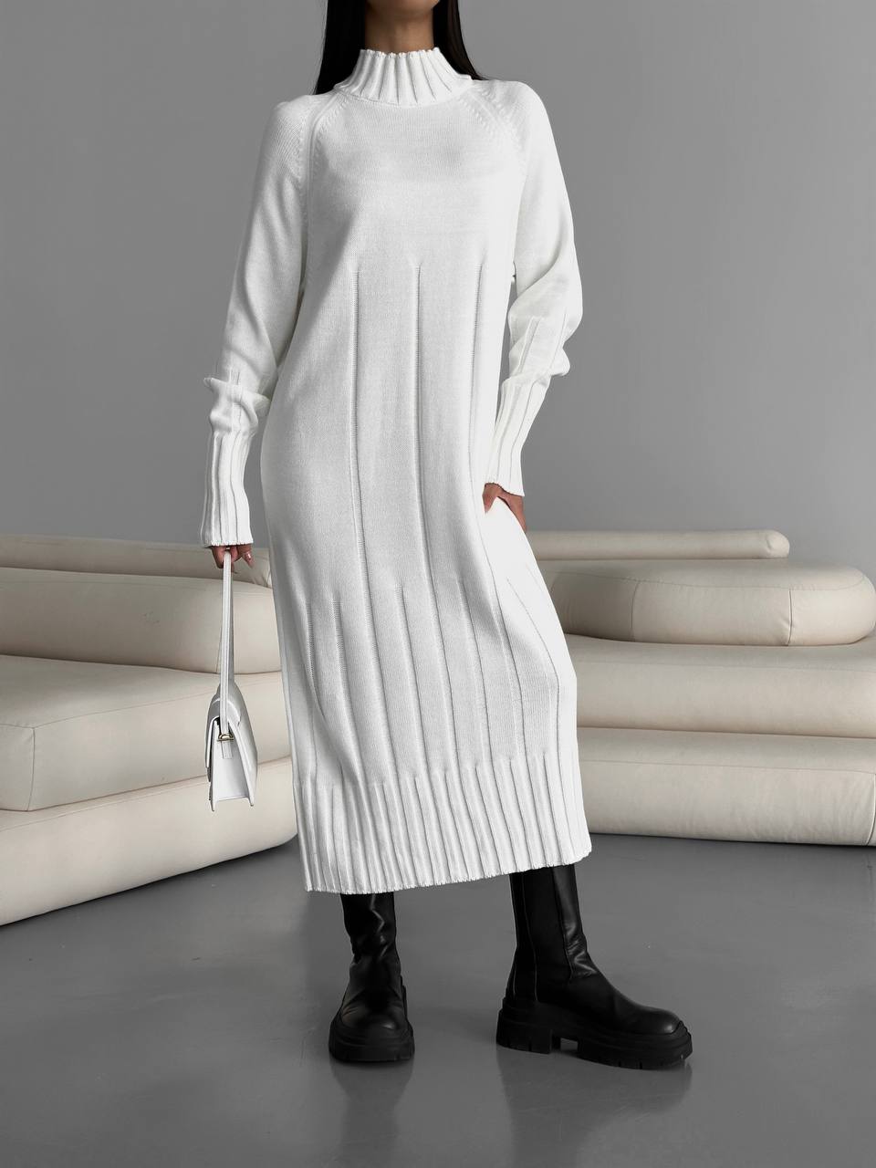Person wearing a long white ribbed sweater dress with black boots in a minimalistic setting.
