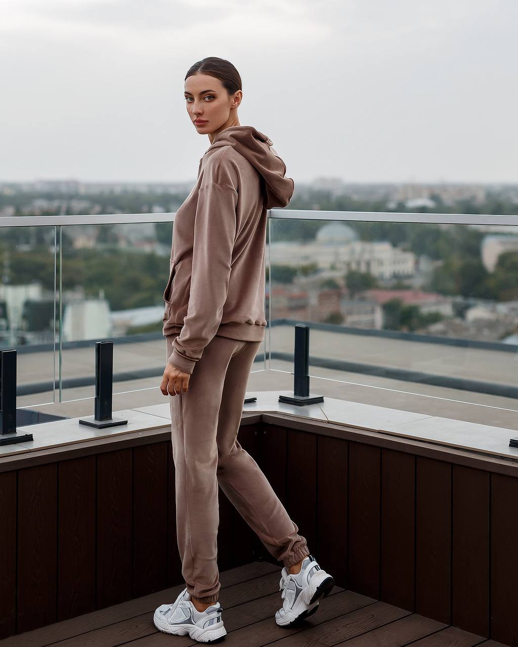Person wearing a beige hoodie and pants on a balcony with a cityscape view.
