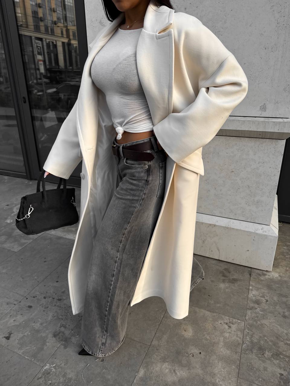 Person wearing a long cream coat, gray top, and jeans outdoors.