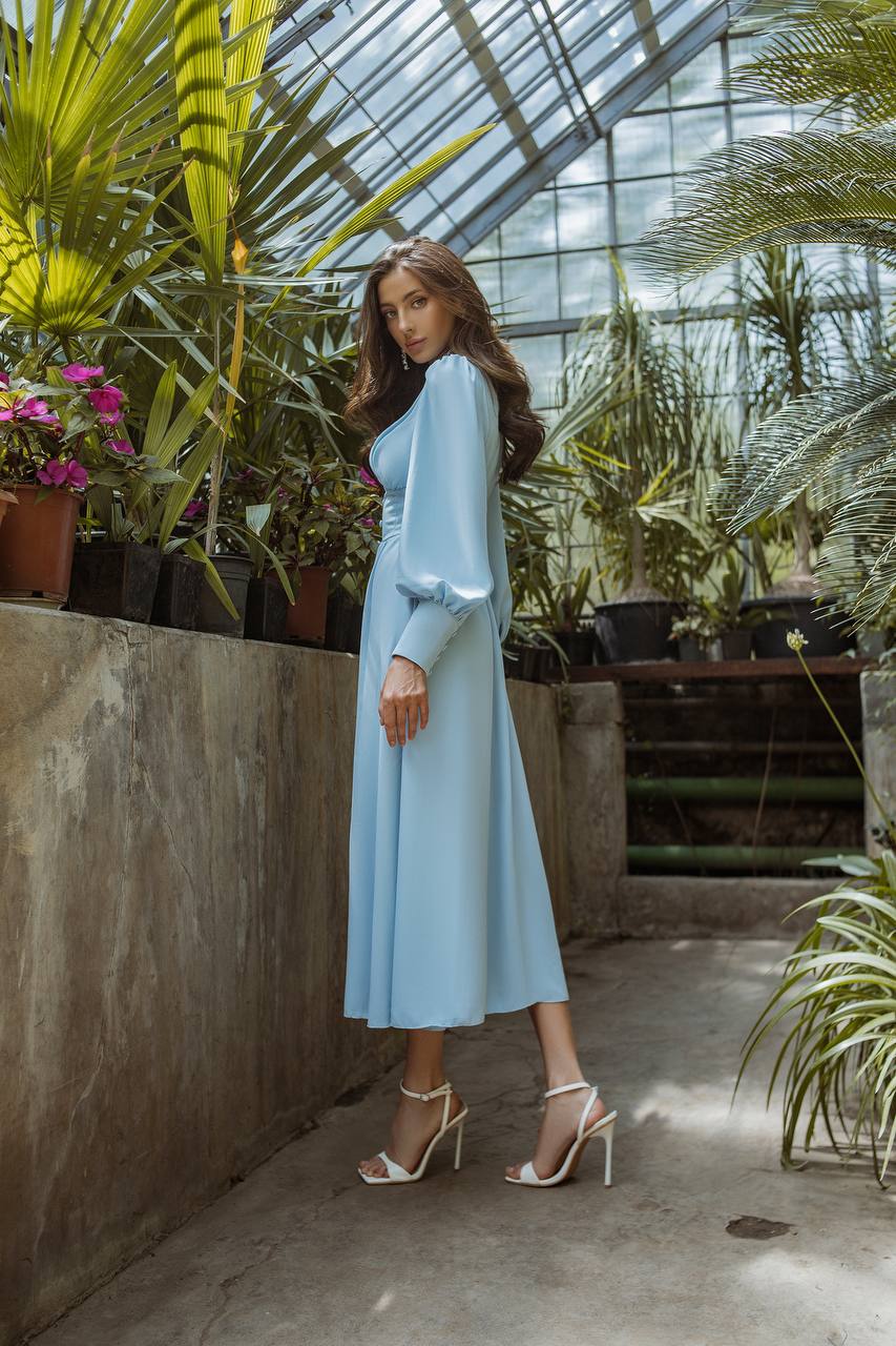 Woman in a light blue dress standing in a greenhouse with plants and flowers.