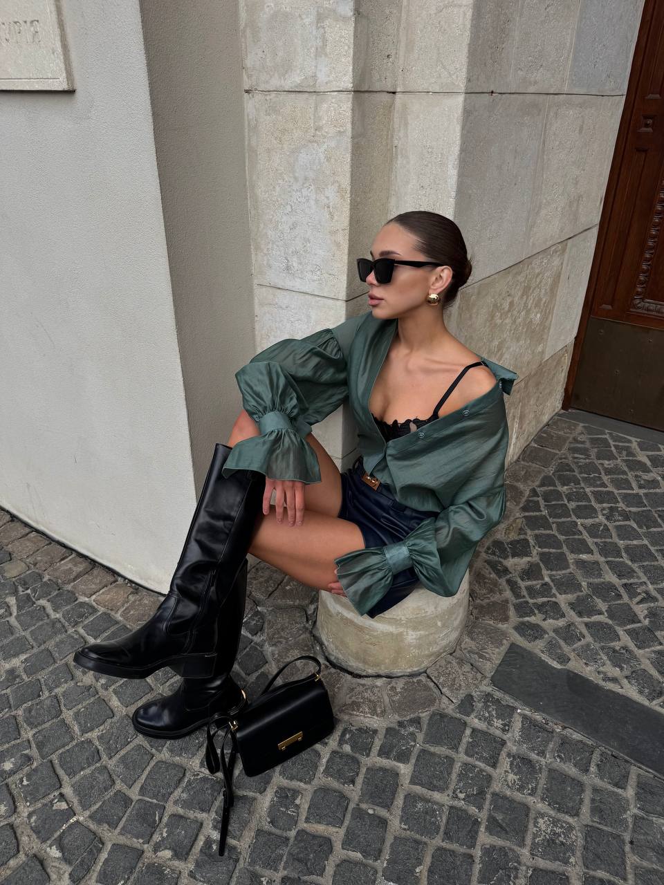 Woman in green shirt and black knee-high boots sitting on a stone bench against a building.