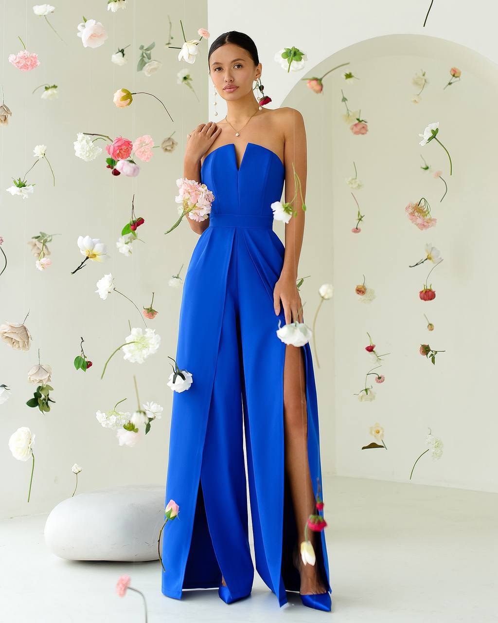 Woman in a blue strapless gown with a slit standing against a floral decorated wall.