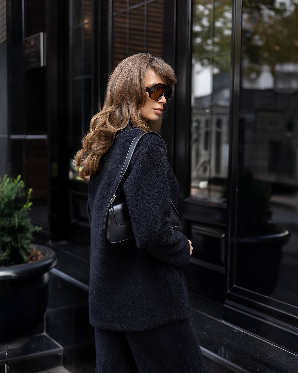 Woman in a dark coat and sunglasses walking past a building entrance.