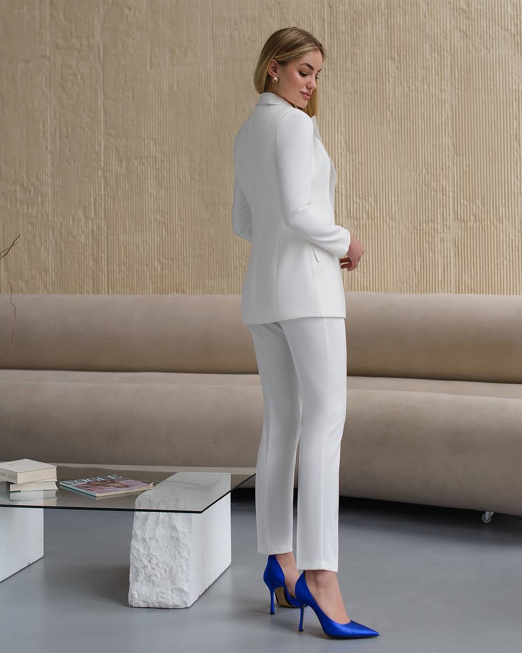Woman in a white suit with blue shoes standing in a modern living room.