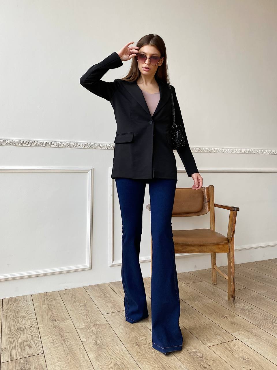 Woman wearing a black blazer and blue flared pants in a room with wooden floor and white walls.