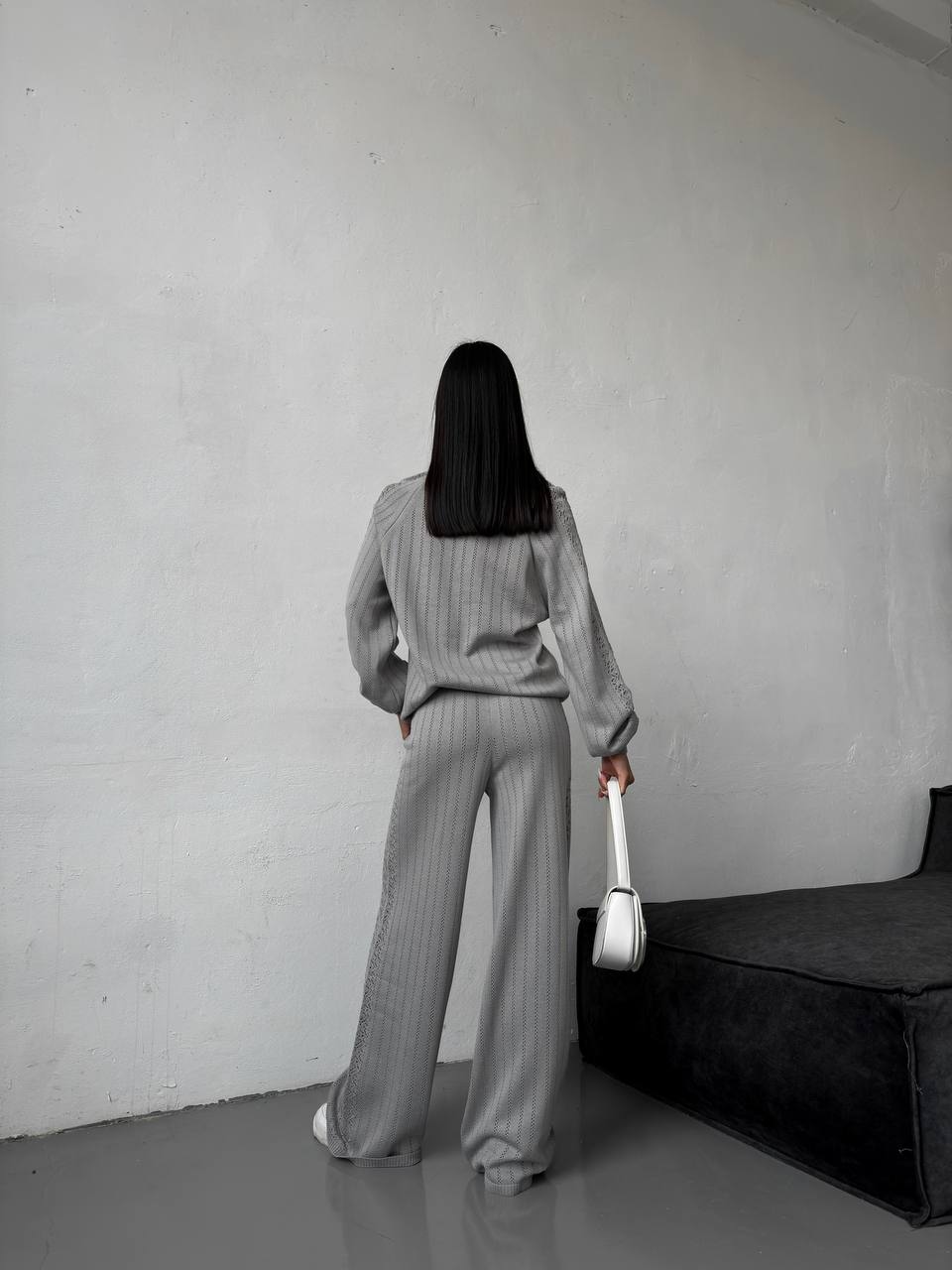 Person wearing a gray outfit holding a white bag against a plain wall.