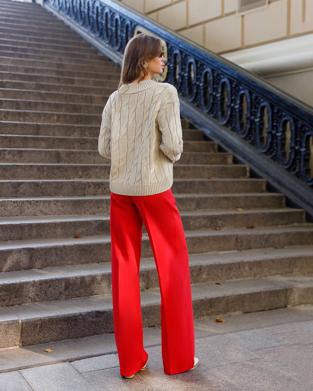 Woman in a beige sweater and red pants standing on stone steps.