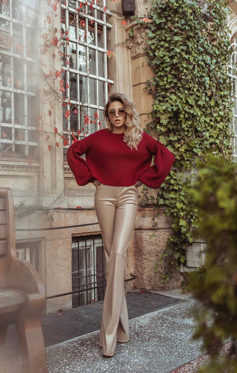 Woman in a red sweater and gold pants standing in an outdoor setting with greenery.