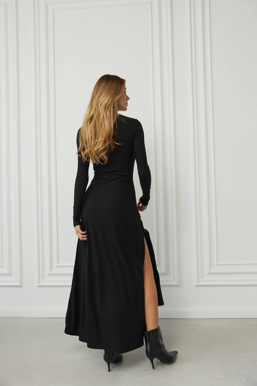 Woman wearing a black dress with a high slit, standing against a white paneled wall.
