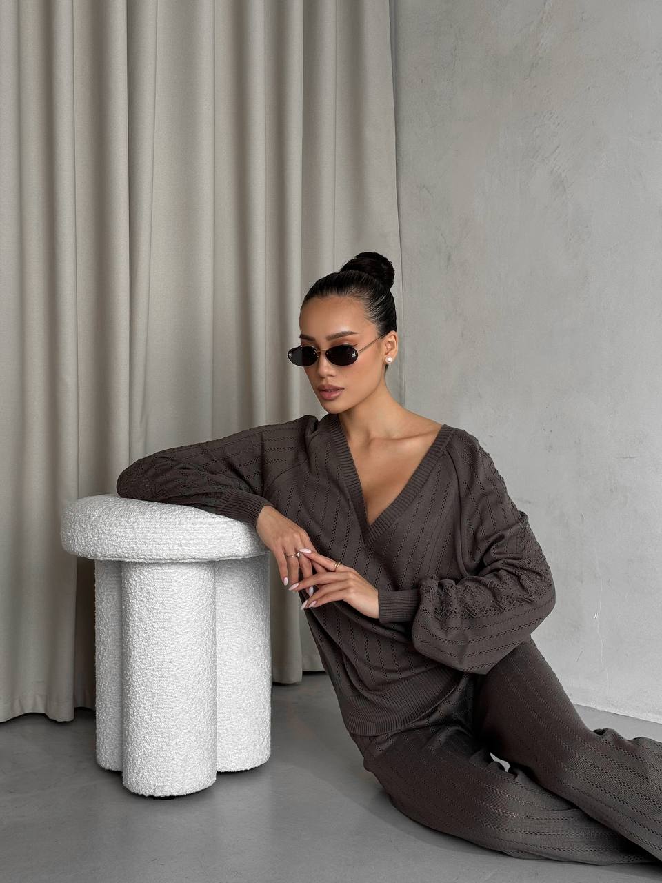 Woman in a brown outfit sitting on a textured white stool against a neutral background
