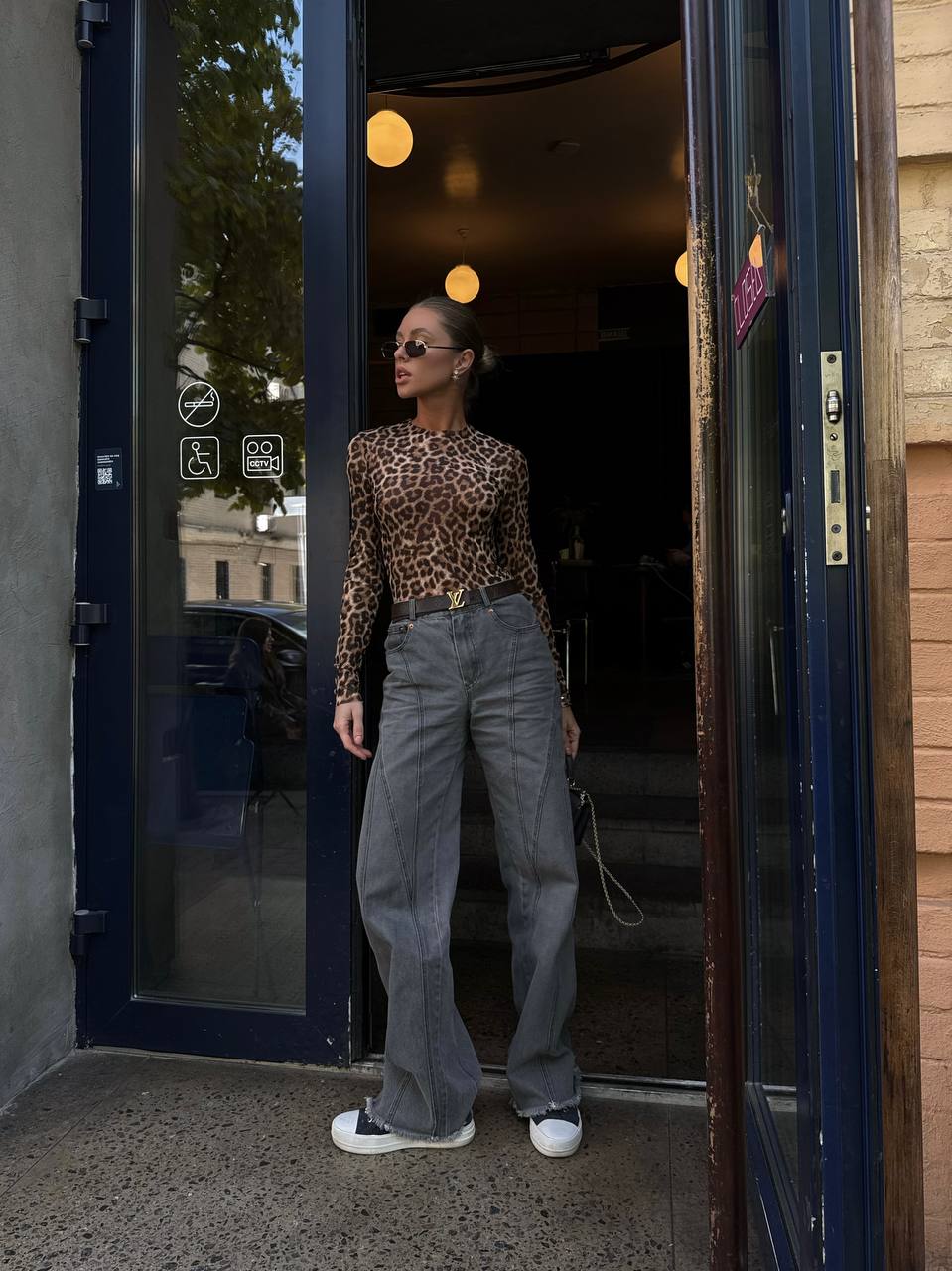 Person wearing a leopard print top and gray pants standing in an open doorway.
