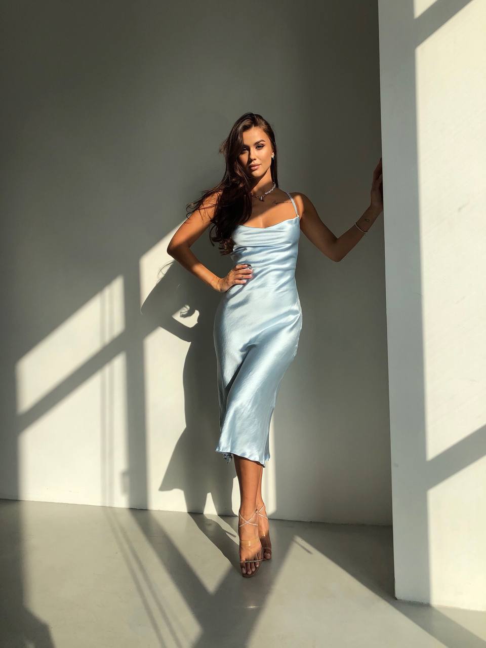 Woman in a light blue dress standing against a gray wall with sunlight casting shadows.