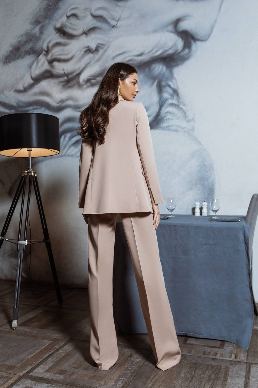 Woman in a beige suit standing in a stylish room with a lamp and table.