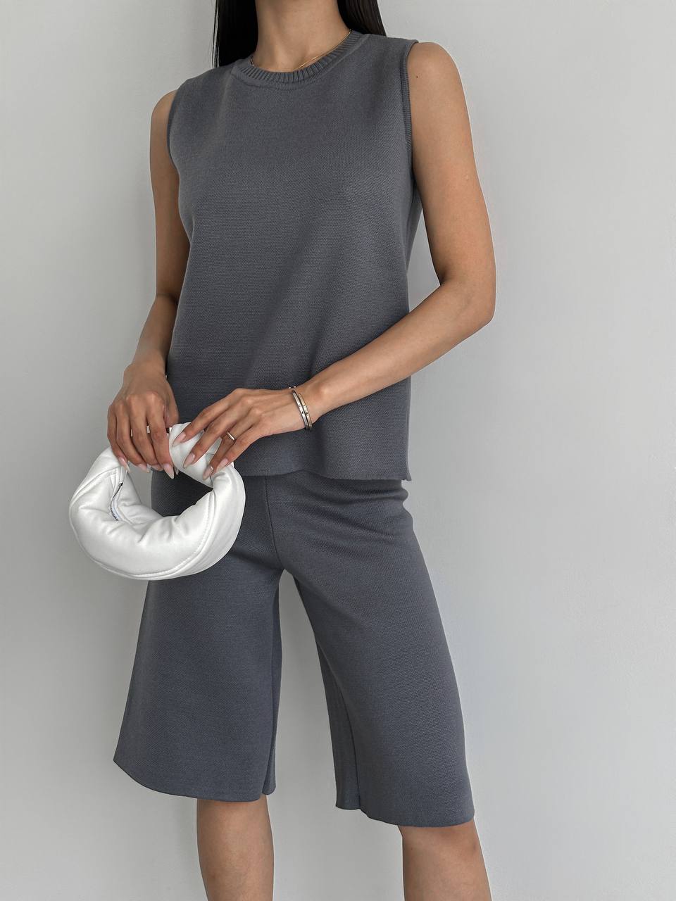 Person wearing a gray sleeveless top and shorts holding a white circular clutch against a plain background