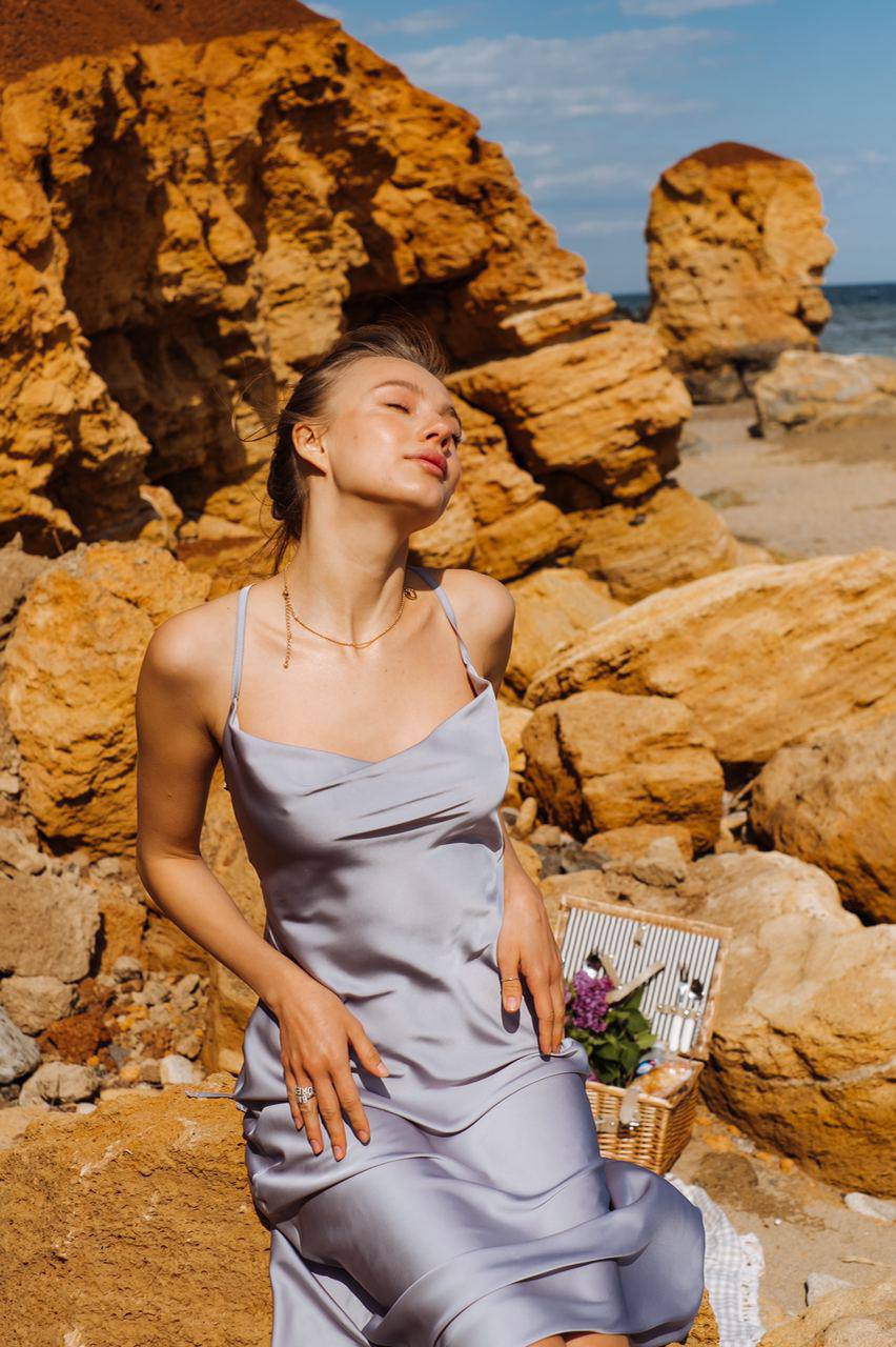 Woman in a light blue dress sitting on a rocky beach with a picnic basket and flowers.