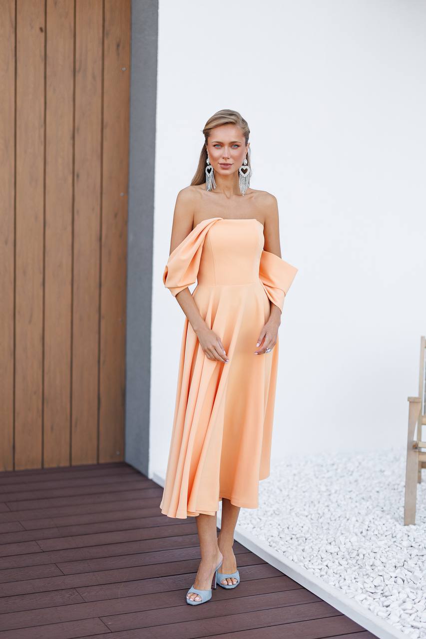 Woman wearing a peach strapless dress standing on a wooden deck with a white wall background.