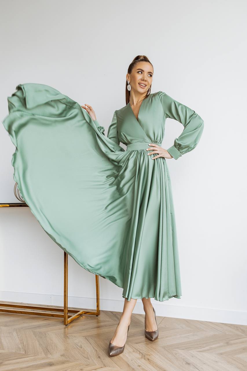 Woman wearing a green dress holding it out in a room with a white wall and wooden floor.