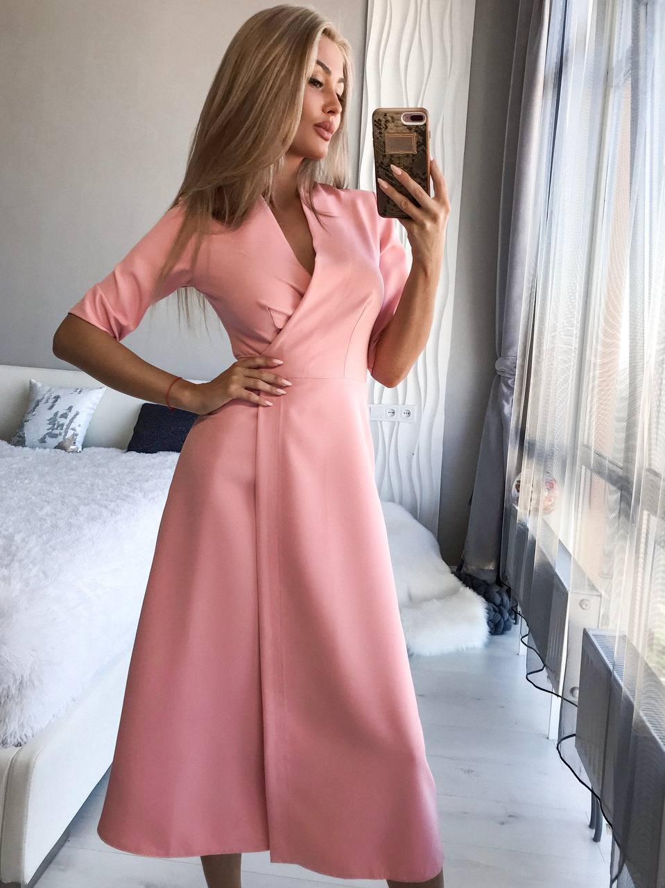Woman in a pink dress taking a mirror selfie in a room with a couch and window.