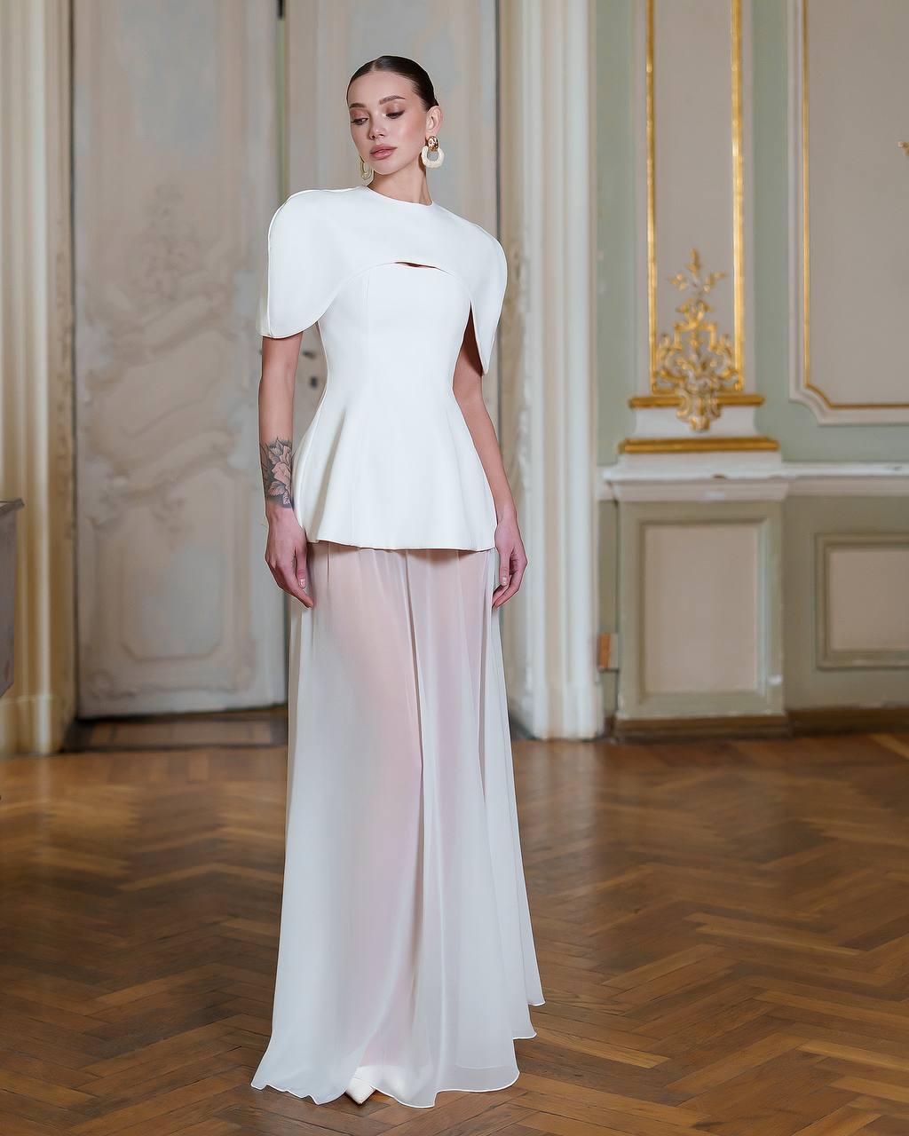 Woman wearing a white peplum top and flowing white skirt in an elegant room with wooden floor and decorative walls.