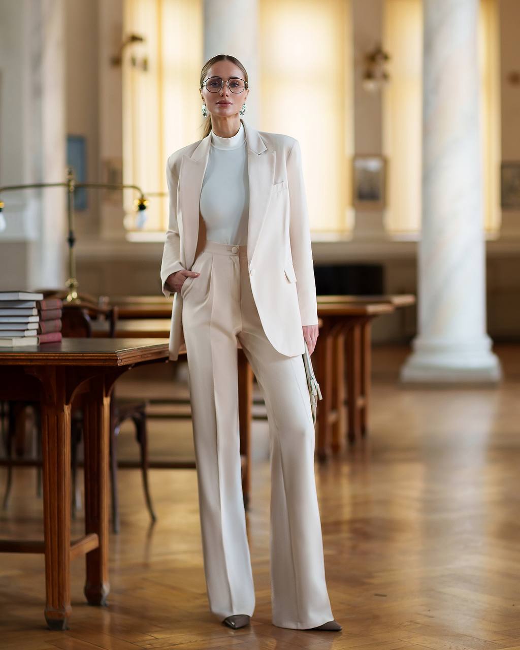 Woman in a beige suit standing in an elegant room with wooden tables and columns.