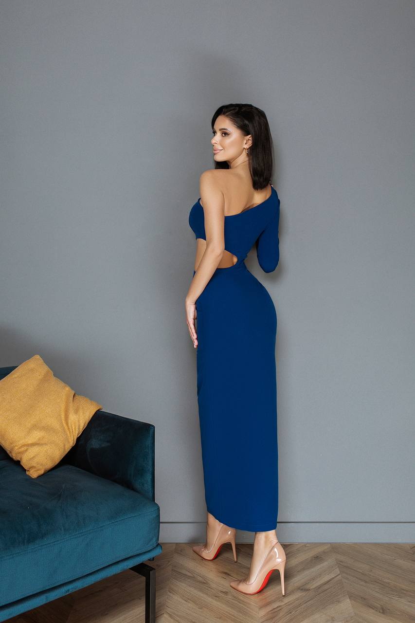 Woman in a blue dress standing against a gray wall with a teal couch and yellow pillow.
