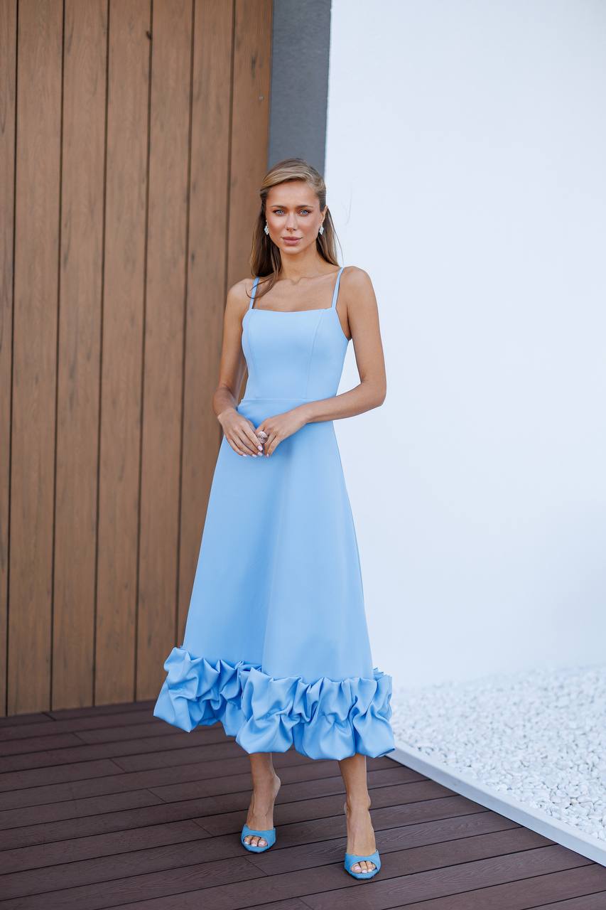 Woman wearing a light blue dress with ruffled hem standing against a wooden and white wall.