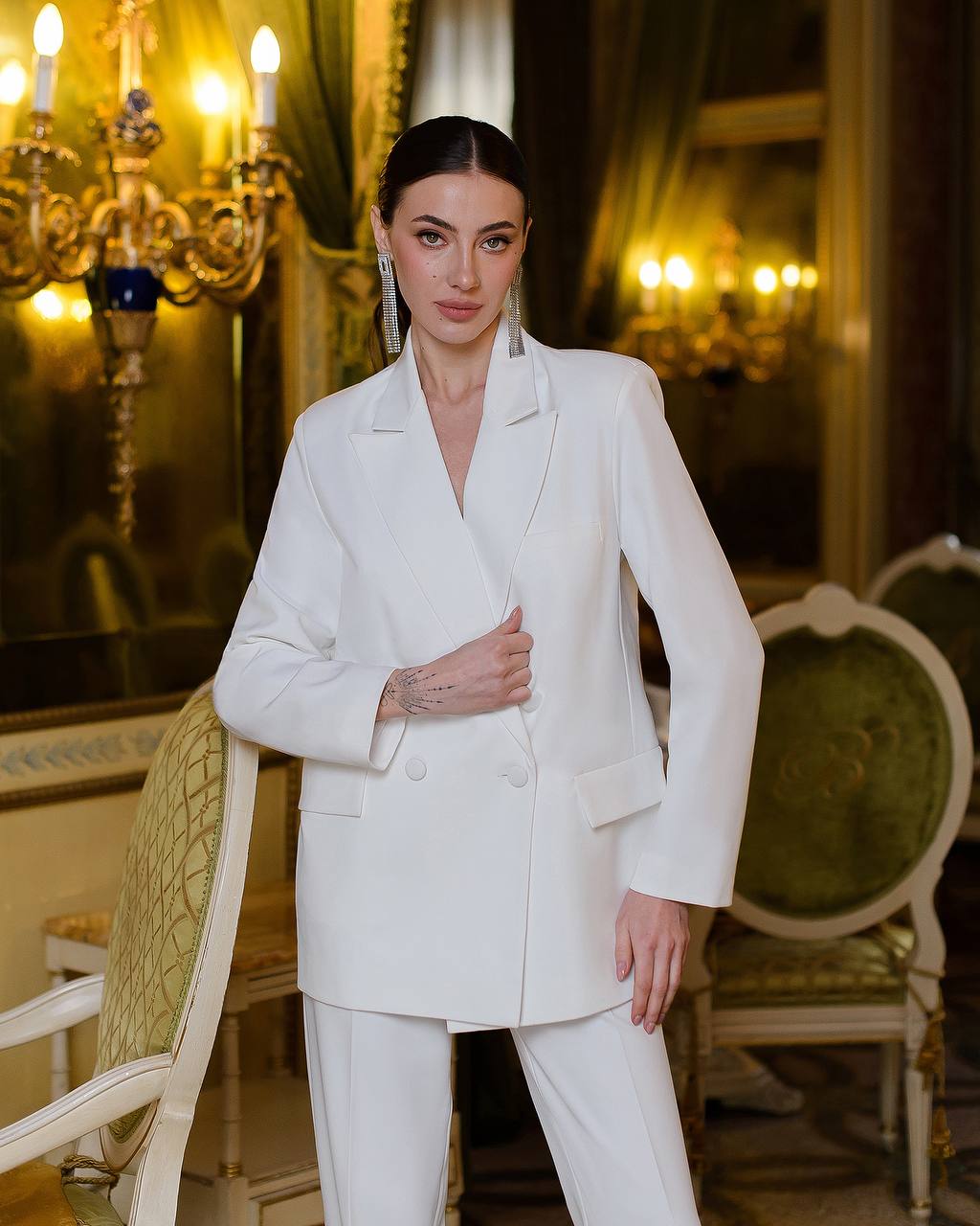Woman in a white suit standing in an elegant room with chandeliers.