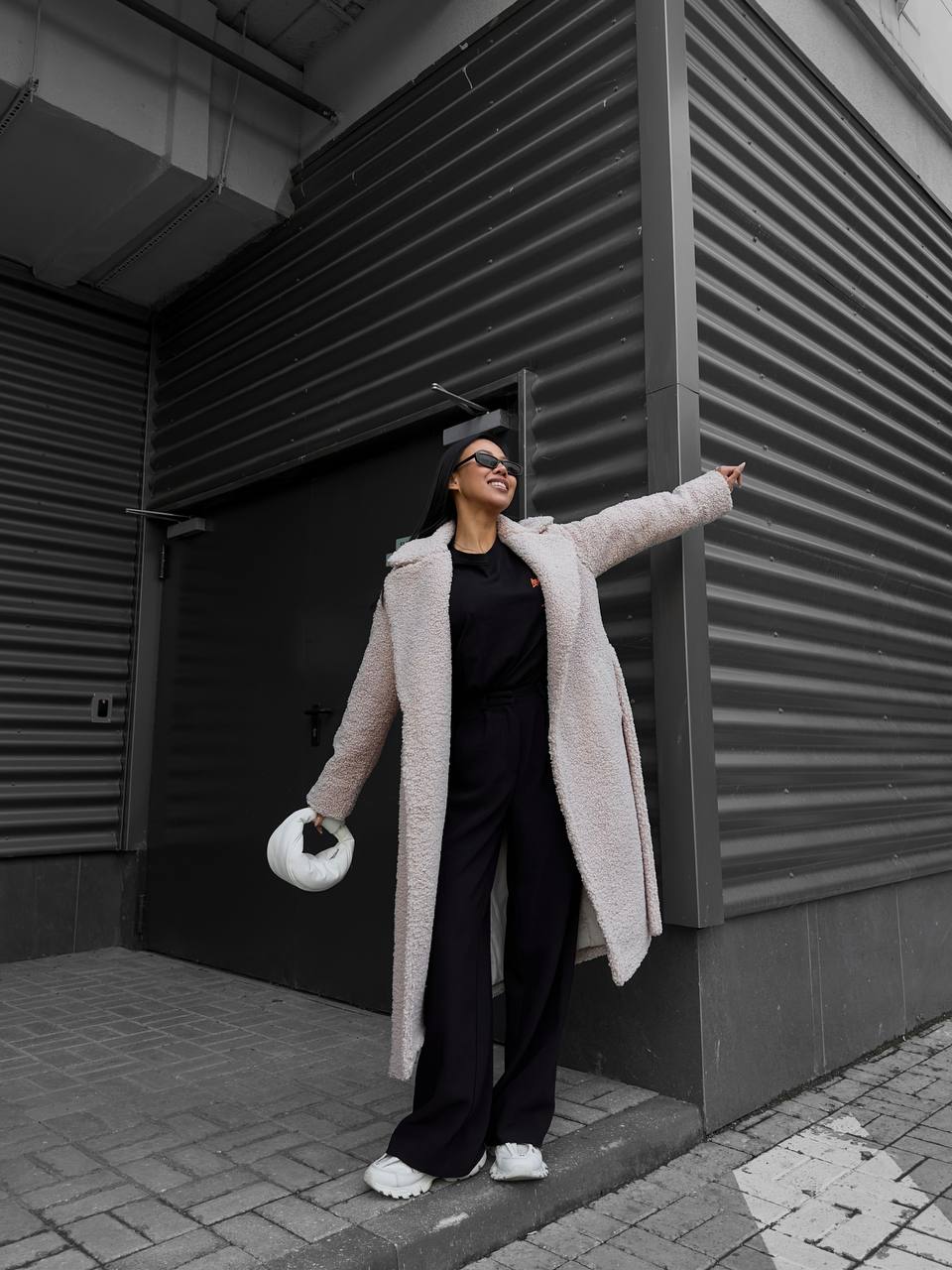 Person wearing a long light pink coat standing against a dark building wall.