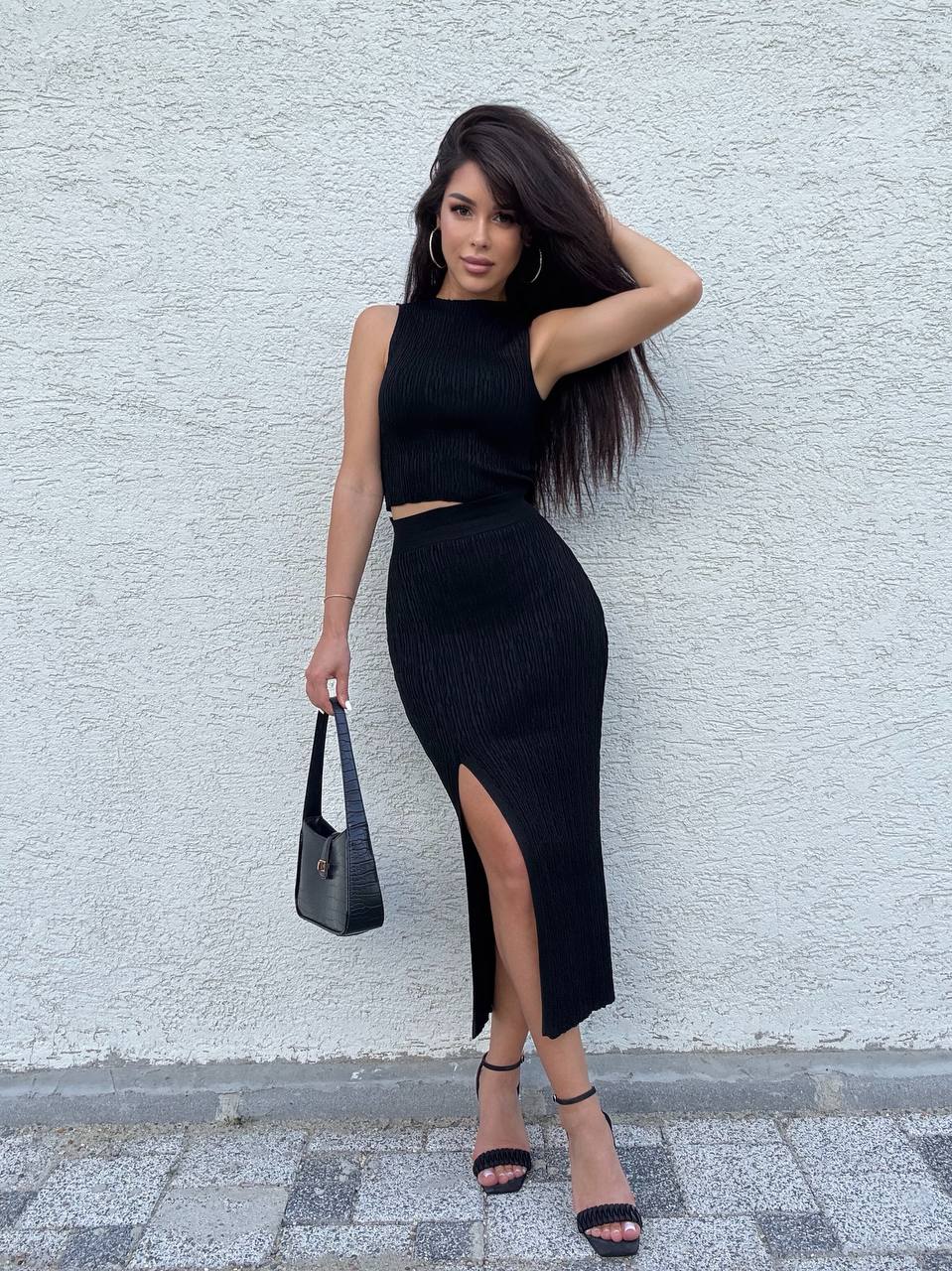 Woman in a black outfit with a slit skirt against a light gray wall.