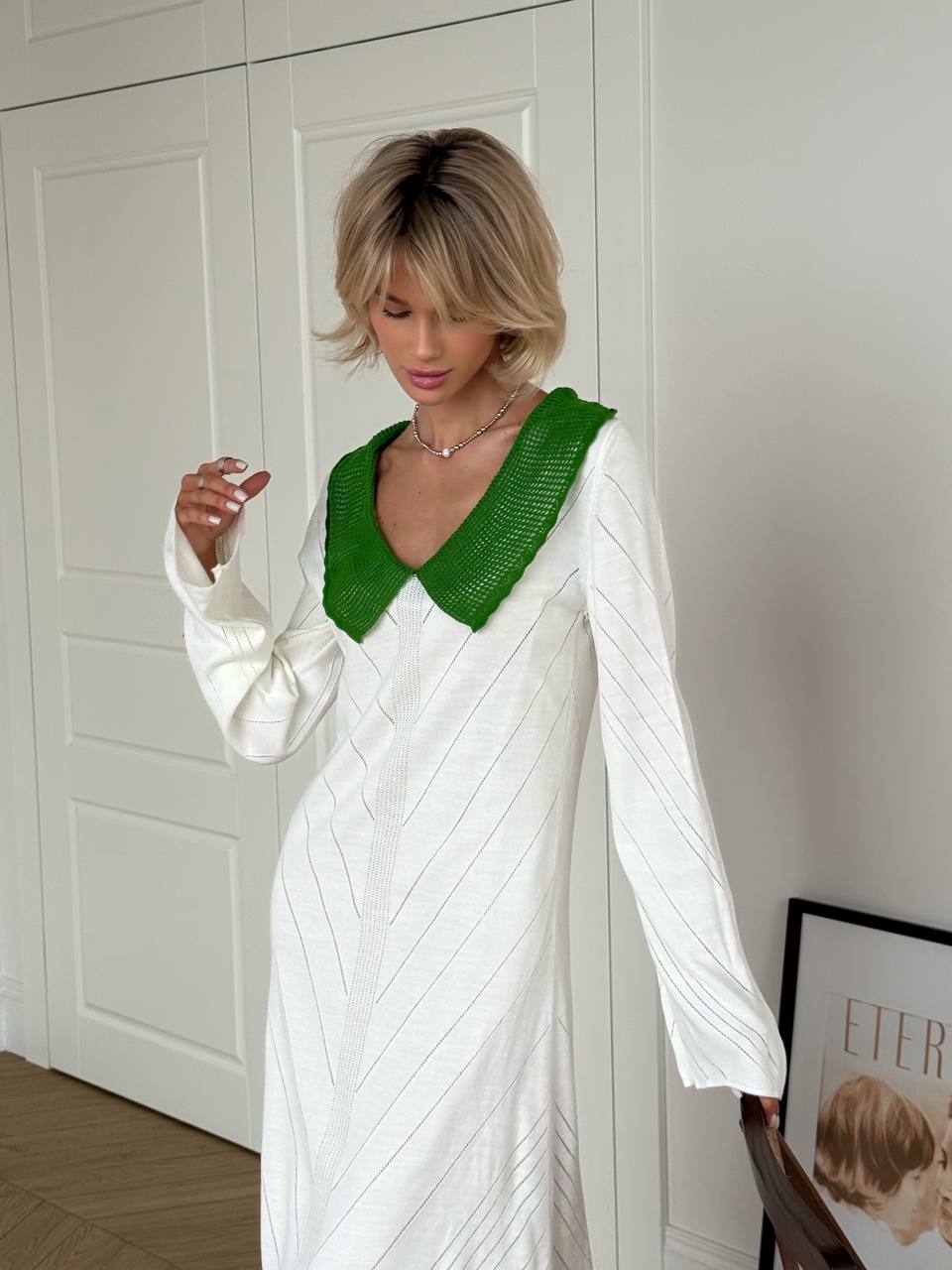 Woman wearing a white dress with a green neckline against a white wall.