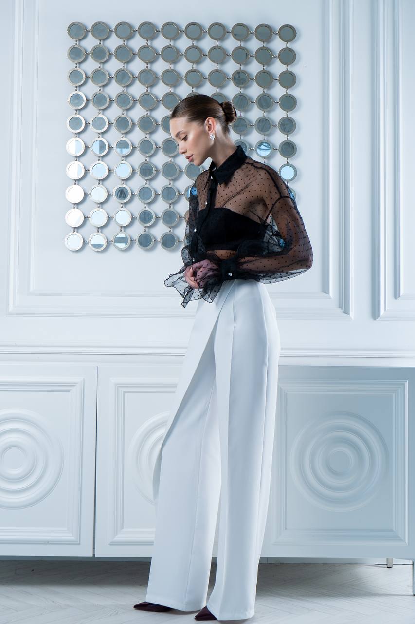 Woman in a stylish outfit with a black top and white pants against a decorative wall.