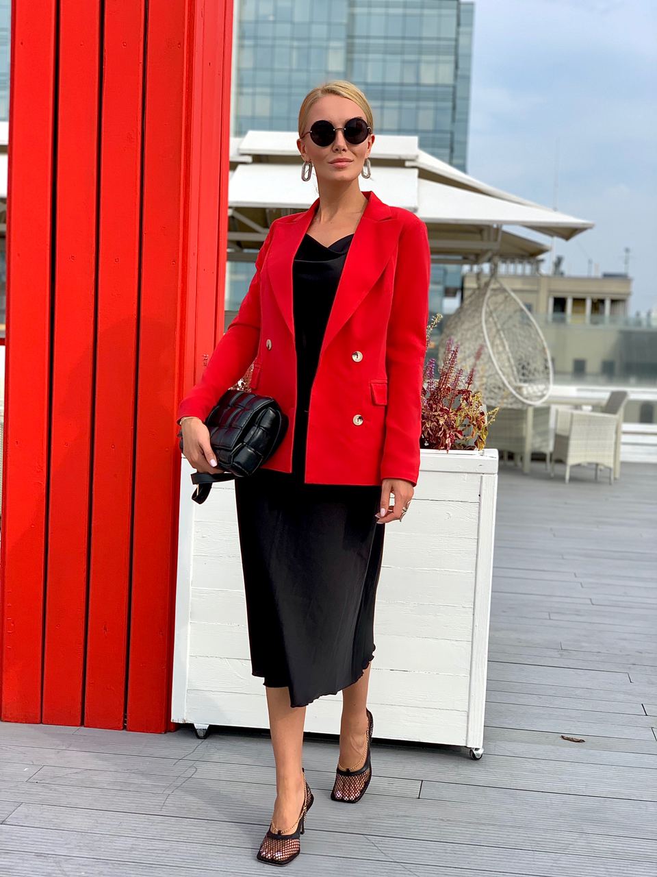 Woman in a red blazer and black outfit standing on a rooftop with a red wall and building in the background.